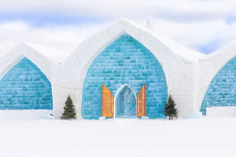 The façade of the Hôtel de Glace (Ice Hotel) near Quebec City, featuring dramatic Gothic-arched walls built from blue ice blocks, wooden entry doors flanked by small evergreen trees, and a snow-covered foreground under a pale winter sky.