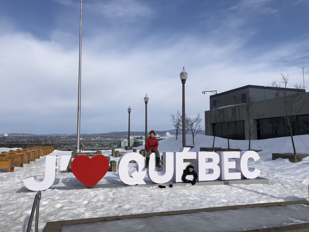 Two children play on large snowy letters that spell "J ❤ QUEBEC" at an outdoor viewpoint in Quebec City in winter. One child in a red jacket sits on top of the letters while another in a dark jacket and green hat crouches in the snow beside them with a wide view of the city and winter sky in the background.