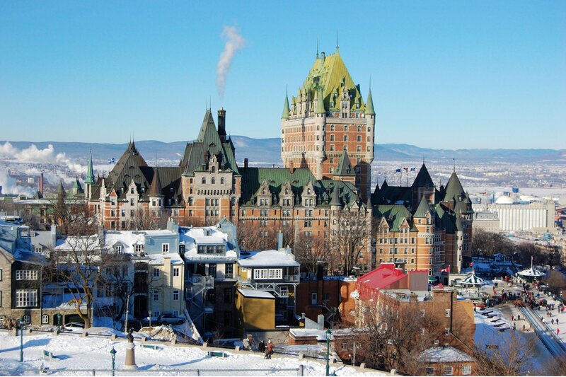 A winter view of Old Quebec City with the iconic Château Frontenac castle-hotel dominating the skyline, its copper-green turrets rising above snow-dusted historic stone buildings, with the frozen St. Lawrence River and distant mountains visible in the background.