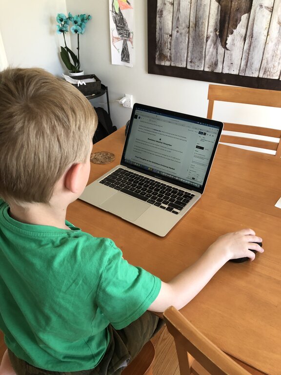 A child in a green t-shirt looks at a laptop screen showing a blog editing platform. A large wooden world map hangs on the wall behind the table.