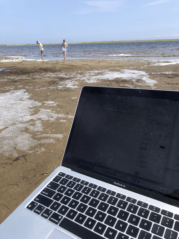 A MacBook Air sits open on a sandy beach with the screen faintly showing a blog interface while two children play near the shoreline in the background. The scene shows a blend of work and family time during travel.