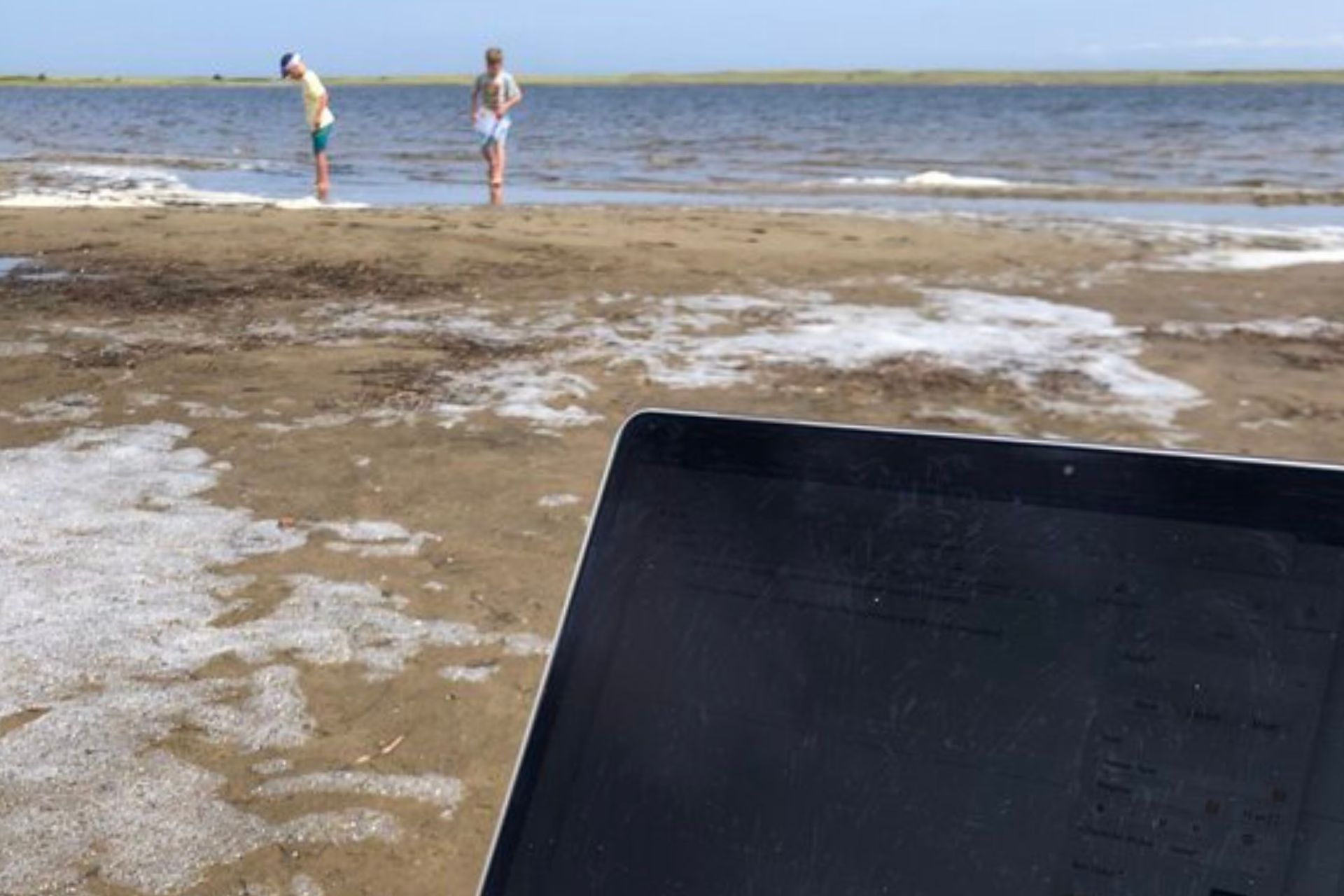 A laptop sits open on a sandy beach while two children play near the shoreline in the background. The photo illustrates the balance of family travel blogging and work alongside family time outdoors.