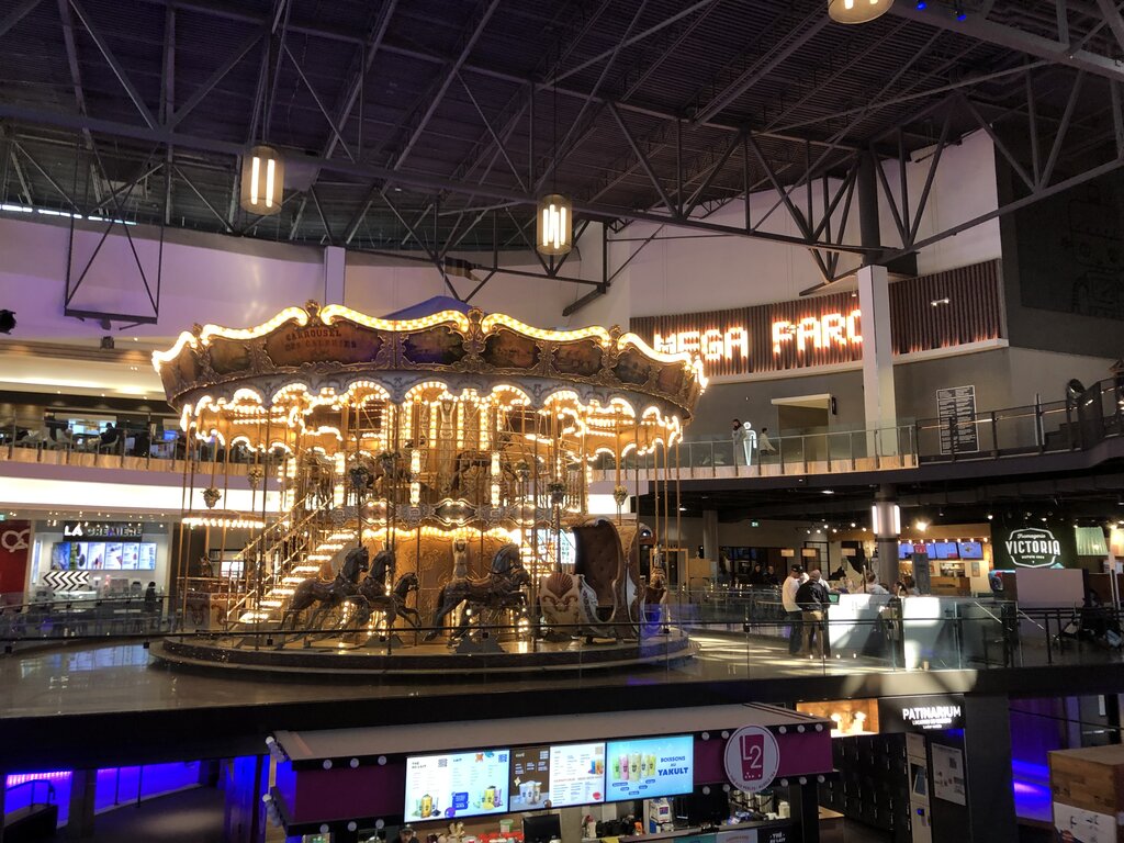 A glowing double-decker carousel lit with golden lights stands at the center of Méga Parc in Quebec City. The vintage-style ride contrasts with the modern shopping center around it, creating a whimsical centerpiece.