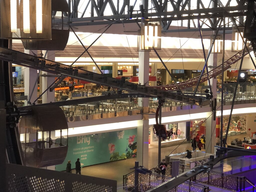 A close-up of the seated zipline at Méga Parc in Quebec City, weaving through the food court and shopping area with visible carts and tracks. The dramatic design adds a thrill to this indoor theme park experience.