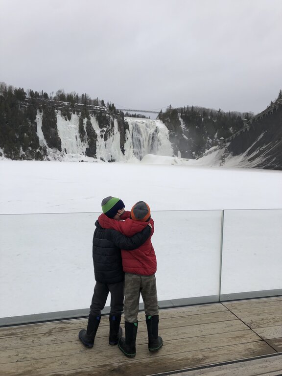 Two children wearing winter coats and boots share a hug on a snowy overlook, facing the partially frozen Montmorency Falls. A scenic and heartwarming moment during a visit to Montmorency Falls in the winter.