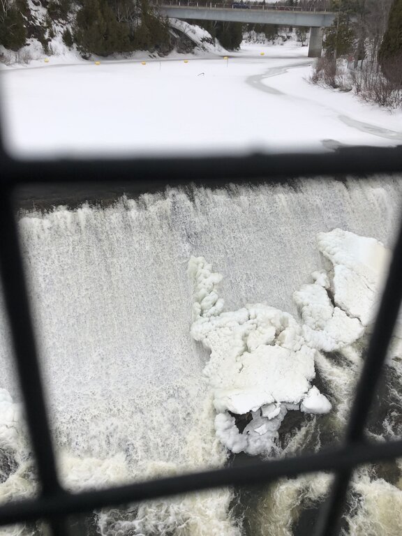 A dramatic top-down view of Montmorency Falls shows foaming whitewater and large frozen ice formations near the river’s edge, framed by a metal fence. This intense perspective captures the power of Montmorency Falls in the winter.