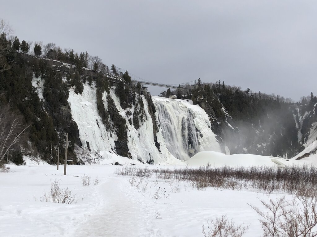 A snowy path leads to Montmorency Falls, with frosty cliffs and a suspension bridge visible above the thundering waterfall. This peaceful trail highlights the natural wonder of Montmorency Falls in the winter.