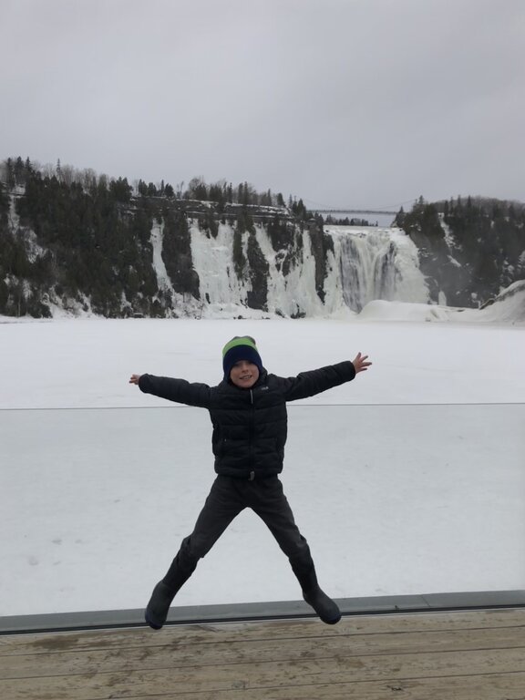 A child dressed in winter clothing jumps joyfully in front of the frozen Montmorency Falls, with snow-covered cliffs and trees in the background. This energetic scene captures the magic of Montmorency Falls in the winter.