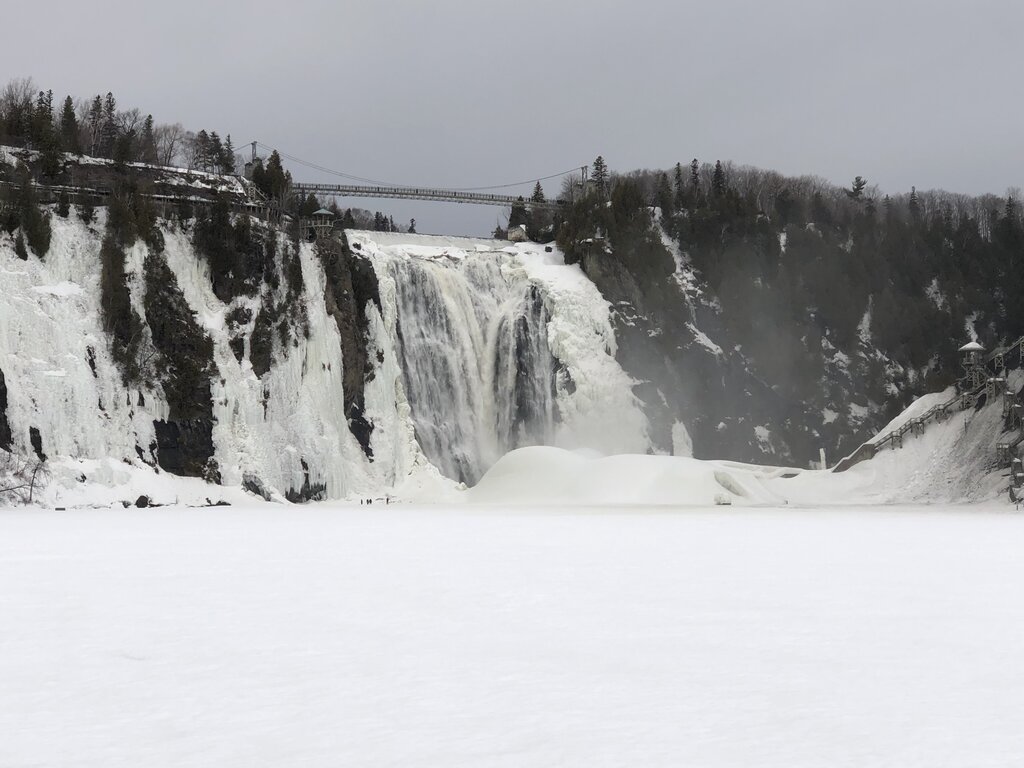 A close-up of Montmorency Falls shows powerful water cascading over icy cliffs, with mist rising and snow blanketing the frozen river below. This dramatic view showcases the raw beauty of Montmorency Falls in the winter.