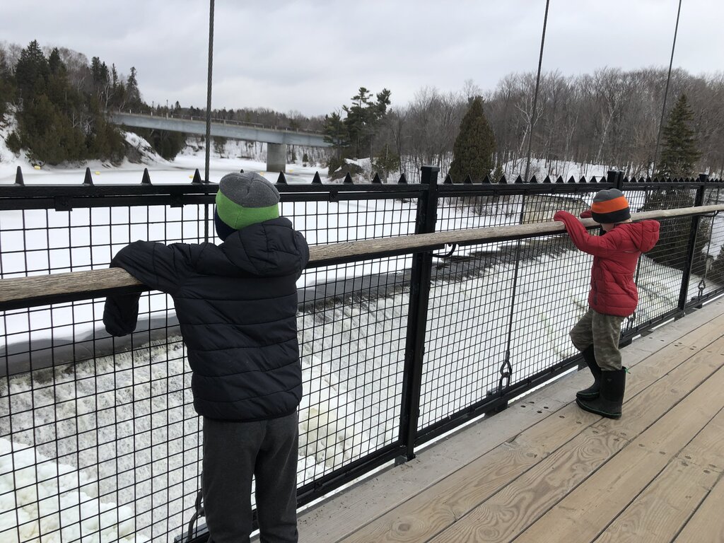 Two children in winter jackets and boots stand on a wooden pedestrian bridge, looking down at a partially frozen river near Montmorency Falls. Snow-covered trees and a distant road bridge frame the quiet winter landscape.