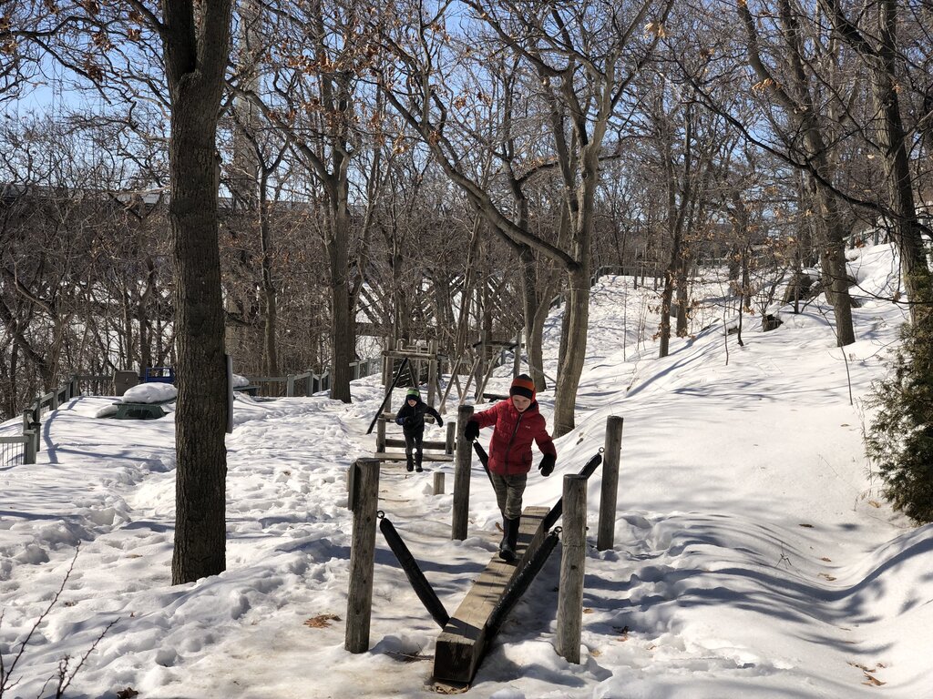 Two children walk across a wooden balance beam in a snowy forest playground, surrounded by leafless trees under a clear blue sky. The scene captures outdoor winter play and adventure on a sunny day in Quebec.