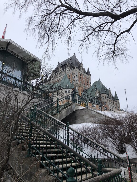 A steep staircase leads up to the Château Frontenac, with ornate green railings and snow-covered trees surrounding the path. The historic hotel overlooks Quebec in winter from its hilltop perch.
