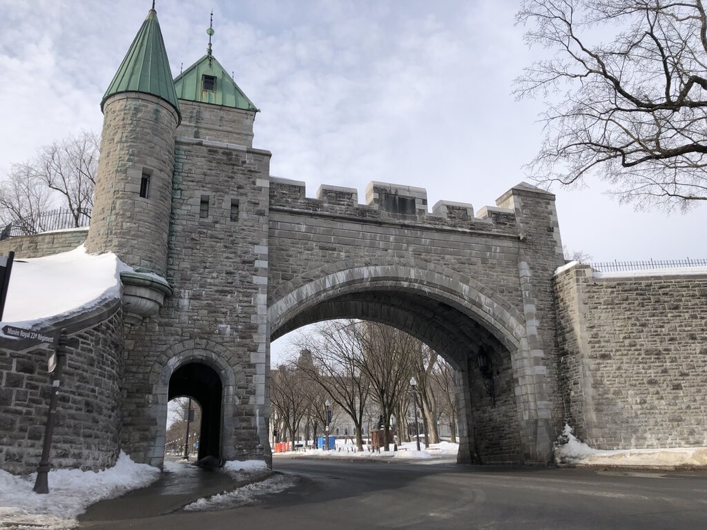A stone arched gate with green-roofed towers, part of Quebec City's fortifications, stands over a snow-dusted road. This historic site is a peaceful winter scene in Quebec.