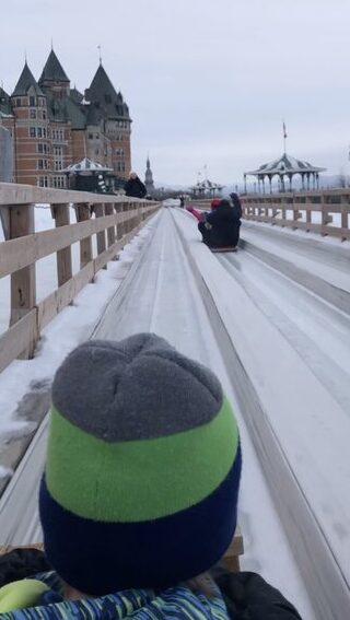 A child rides a wooden toboggan down an icy slide on Terrasse Dufferin in Quebec City, with Château Frontenac and a snow-covered gazebo visible in the background. This thrilling winter activity is a highlight of visiting Quebec in winter.