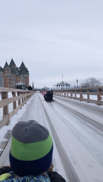 A child rides a wooden toboggan down an icy slide on Terrasse Dufferin in Quebec City, with Château Frontenac and a snow-covered gazebo visible in the background. This thrilling winter activity is a highlight of visiting Quebec in winter.