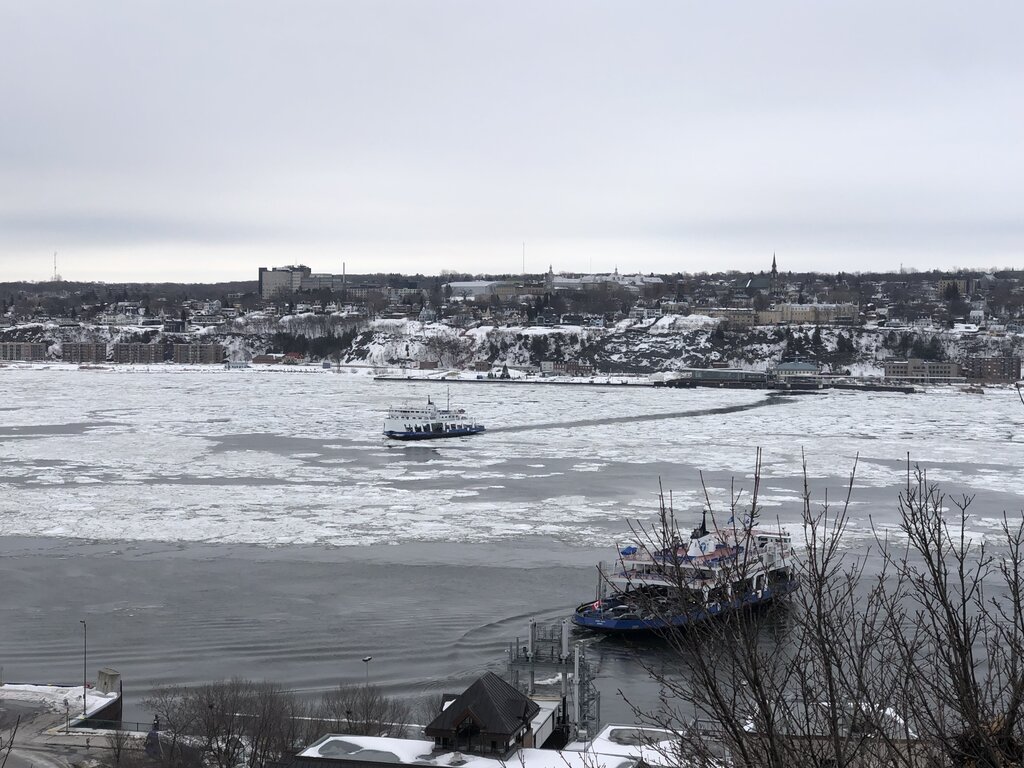 A ferry crosses the partially frozen St. Lawrence River with Lévis visible on the opposite bank, under a gray winter sky. The icy landscape captures the cold, expansive beauty of Quebec in winter.
