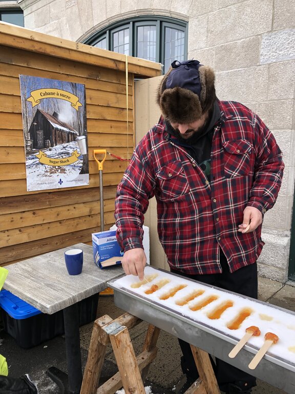 A man in a fur-lined hat and plaid coat prepares maple taffy on snow at an outdoor sugar shack stand, with a "Cabane à sucre" sign on the wall behind him. This traditional maple treat is a highlight of Quebec in winter.
