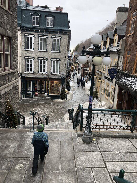 A young boy in a snow suit walks down icy steps toward a cobblestone street in Old Quebec, lined with historic buildings decorated for winter. Snow piles and street lamps add charm to this classic view of Quebec in winter.