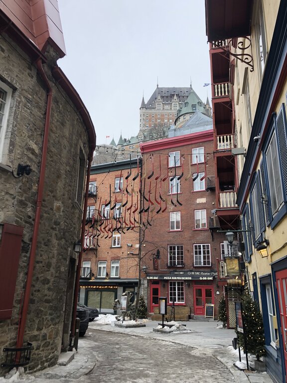 A narrow, snow-lined alley in Old Quebec leads to a red brick building with dozens of red and black hockey sticks hanging above. Château Frontenac towers in the background, adding grandeur to this Quebec in winter setting.
