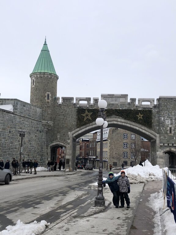 Two children in winter coats stand in front of Porte Saint-Jean, a stone city gate decorated with stars and ivy. This iconic entrance to Old Quebec highlights the historic charm of Quebec in winter.