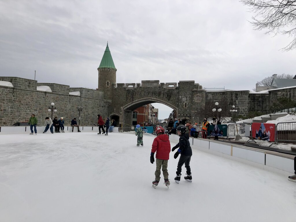 Children and adults ice skate on a public rink near a stone fortress wall and archway in Quebec City. The cloudy sky and snowy surroundings reflect the joy of outdoor activities in Quebec in winter.