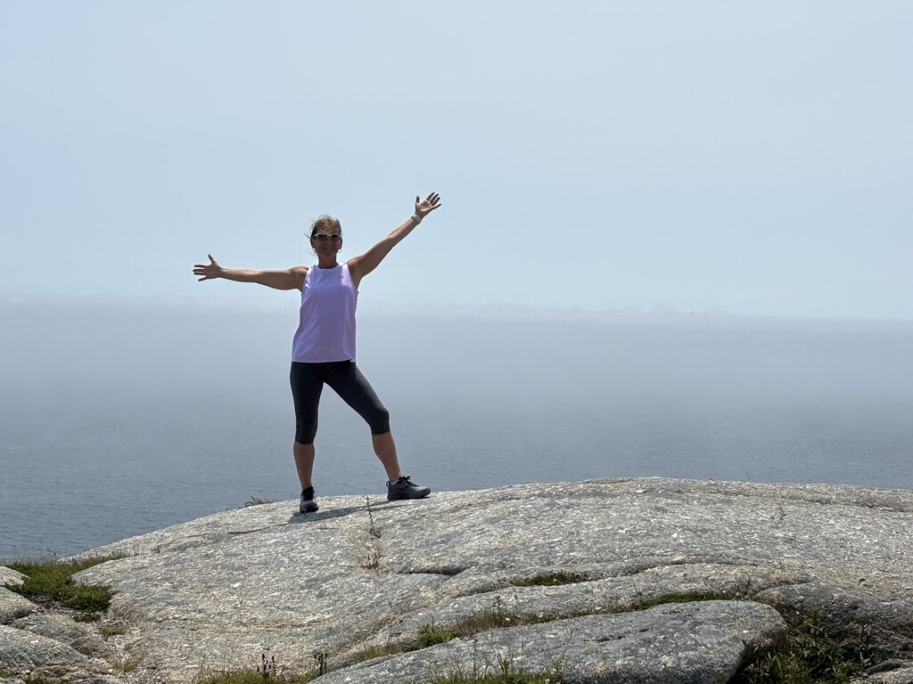 A woman stands on a large, smooth rock with arms outstretched, smiling against a misty ocean backdrop on a hike along the East Coast. The hazy sky and distant horizon blend together, giving a peaceful and expansive feel to the scene.