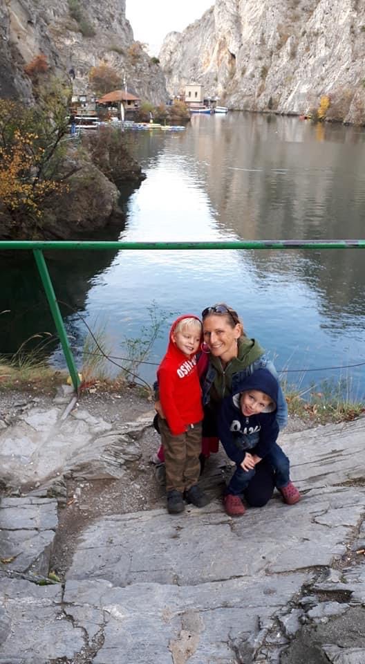A woman crouches beside two young children in hoodies, all smiling on a rocky path overlooking the calm waters of Matka Canyon in North Macedonia. Towering limestone cliffs and autumn-colored trees frame the scenic spot, capturing a moment from one of their adventures together.