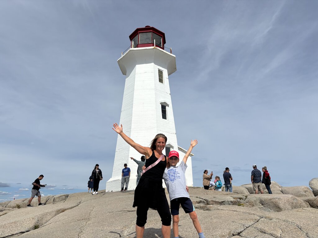 A woman and child pose with arms raised in front of the iconic white lighthouse at Peggy’s Cove, Nova Scotia. They stand on smooth coastal rocks under a clear blue sky, with other visitors exploring the scenic area around them.