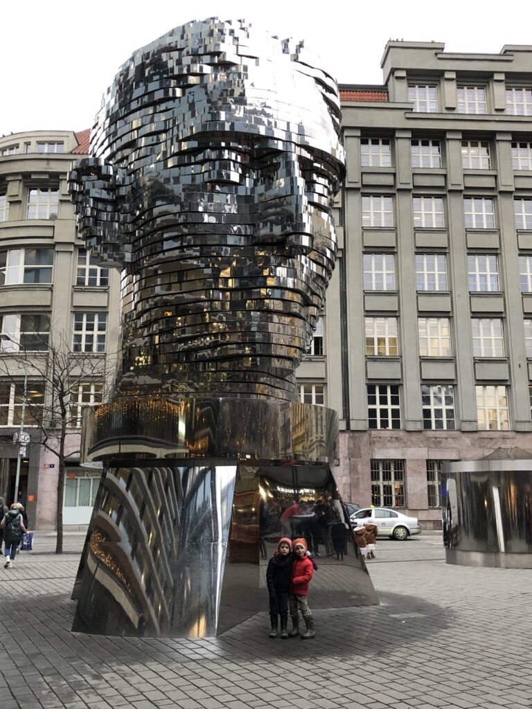 Two children stand at the base of the rotating metallic sculpture of Franz Kafka’s head in Prague, made of reflective horizontal layers that align to form a face. The towering artwork by David Černý is set in a modern urban square, surrounded by gray office buildings and pedestrians.