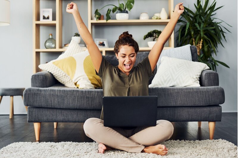 A woman sitting cross-legged on the floor in front of a laptop throws her arms up in excitement, smiling broadly. She’s in a cozy living room with a modern gray couch, decorative pillows, and a wall shelf with plants and decor in the background, creating a casual and celebratory atmosphere.