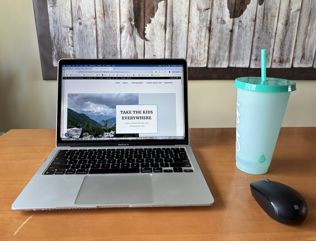A MacBook Air sits open on a wooden desk, displaying a travel blog homepage with the headline “TAKE THE KIDS EVERYWHERE” against a mountain landscape background. Beside the laptop is a large aqua-colored tumbler with a straw and a wireless mouse, all set in front of a rustic wooden wall art piece showing a world map.