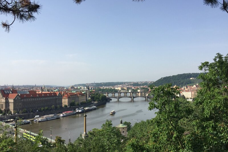 A panoramic view of the Vltava River in Prague, seen from Letna Park, with several historic stone bridges crossing the water, surrounded by red-roofed buildings and green hills.