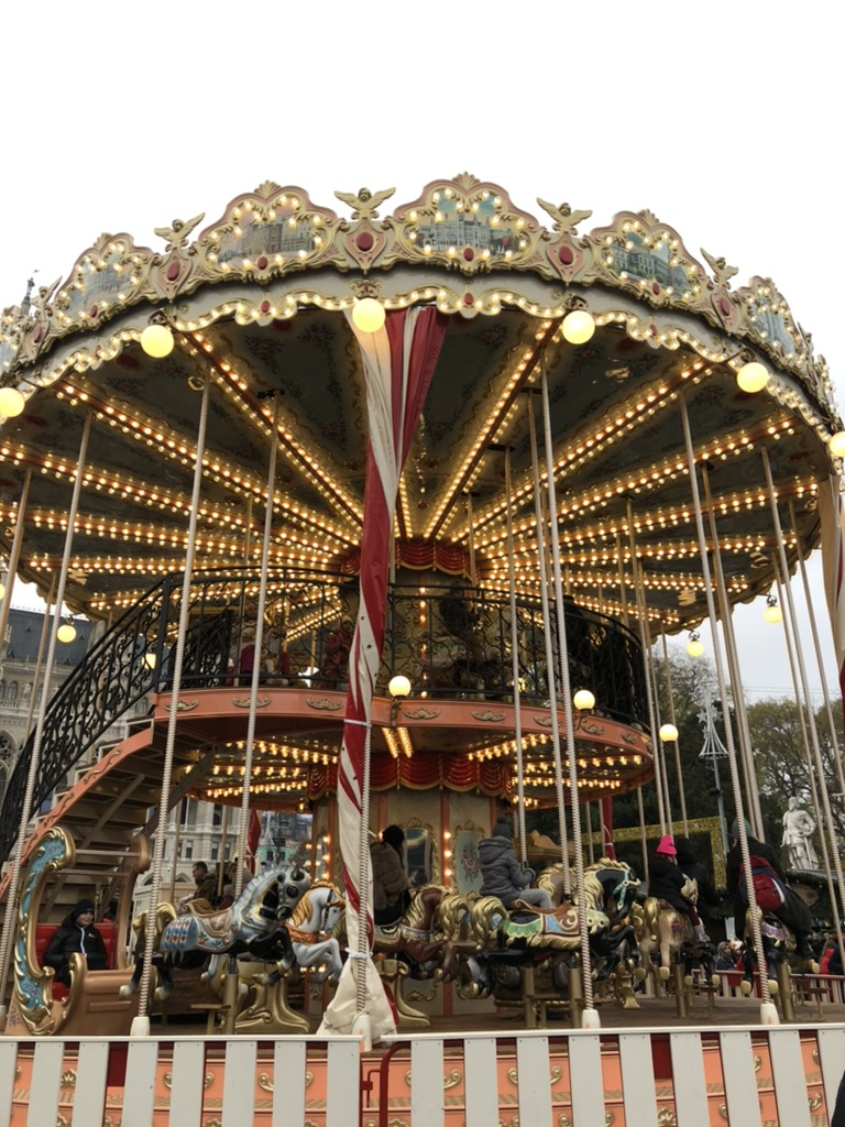 A vintage-style two-story carousel adorned with glowing lights and ornate decorations features painted horses and carriages, with children riding and a spiral staircase leading to the upper level. The scene is set during a cloudy day at Vienna's main christmas market