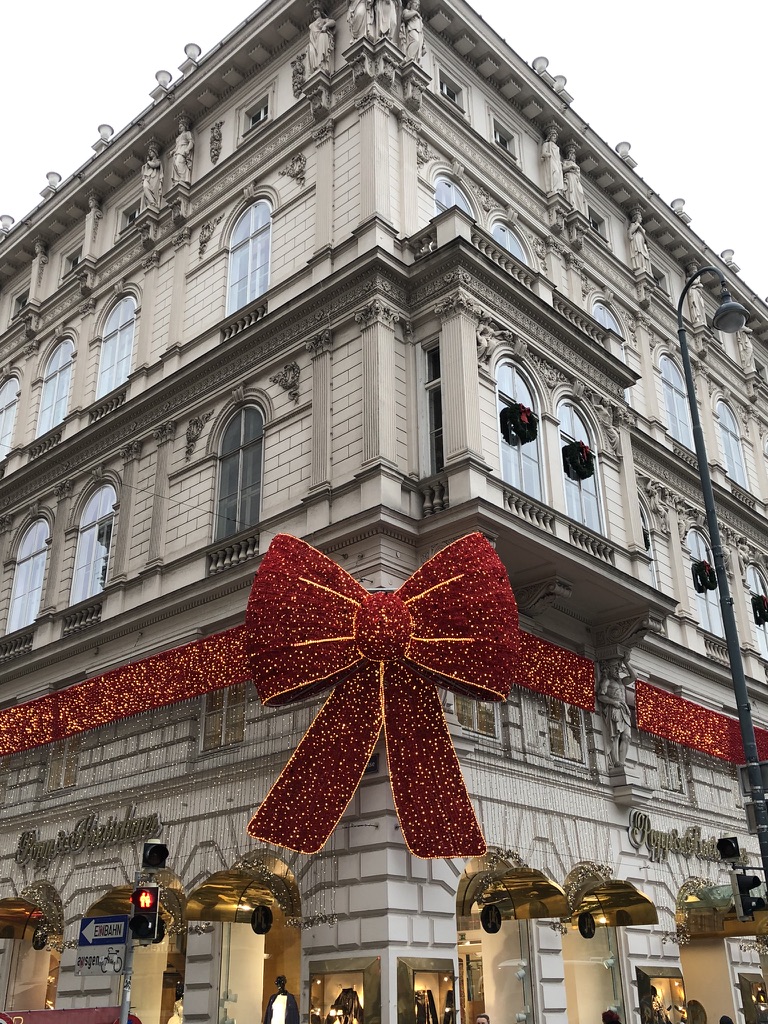 A grand historic building in Vienna is decorated for the holidays with a massive red glittering bow wrapping around its corner, above luxury storefronts with golden awnings. Festive wreaths hang in the windows and classical statues line the rooftop.