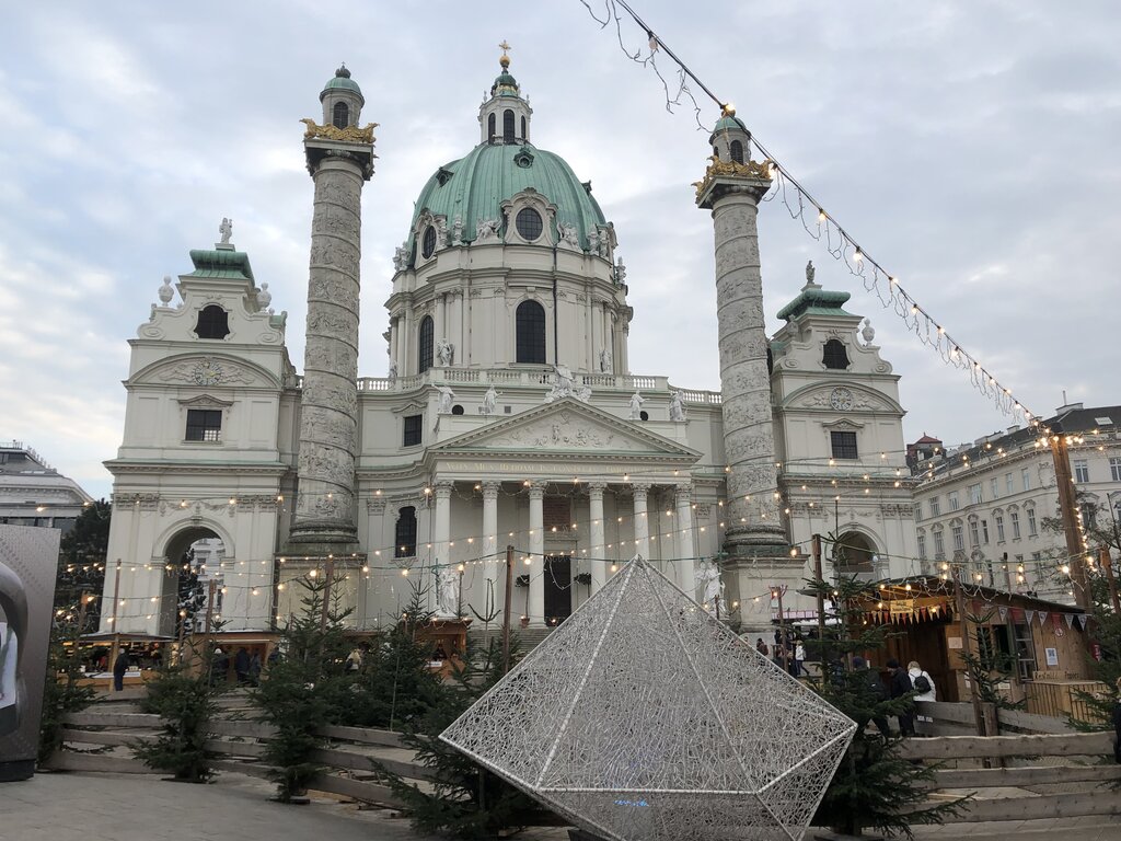 Karlskirche’s baroque façade and twin columns rise behind a Christmas market set up with wooden stalls and string lights, with a geometric wire sculpture and small trees in the foreground during daytime.