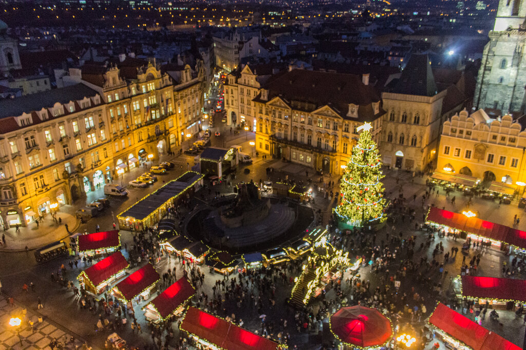 An aerial view of Prague’s Old Town Square Christmas market shows red-roofed stalls, a glowing Christmas tree, and crowds enjoying the festive atmosphere surrounded by historic buildings.