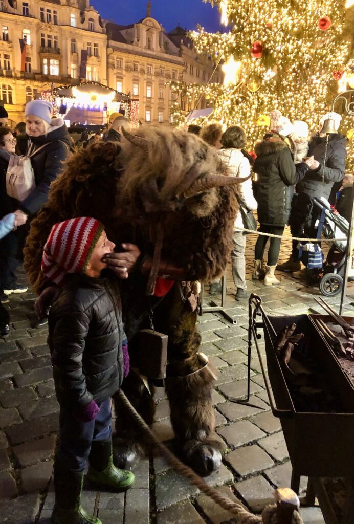 A child in a striped hat smiles as they interact with a person dressed in a furry horned costume at a lively Christmas market surrounded by crowds and lights.