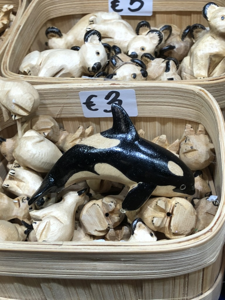 A hand-carved wooden orca painted black and cream rests atop a pile of small animal figurines in a basket marked with a €3 price tag, part of a display of wooden toys at a market stall. The surrounding figures include koalas and other animals with painted details.