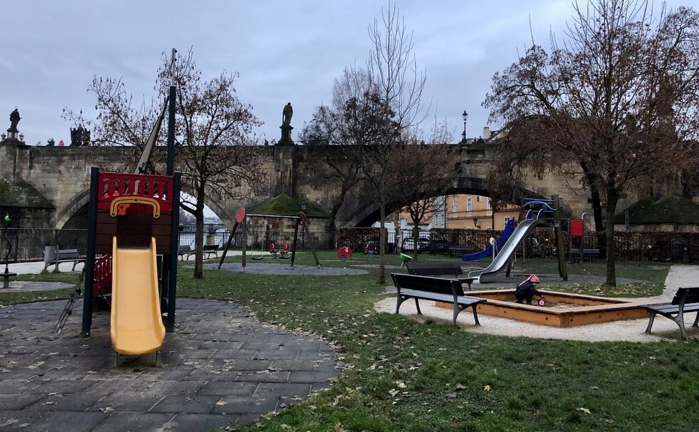 A small playground with slides, swings, and benches sits under the stone arches of the historic Charles bridge, with bare trees and a child playing in a sandbox.