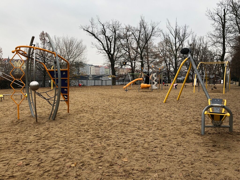 A sandy riverside playground with modern climbing structures and swings, framed by bare trees and colourful city buildings across the water in Prague