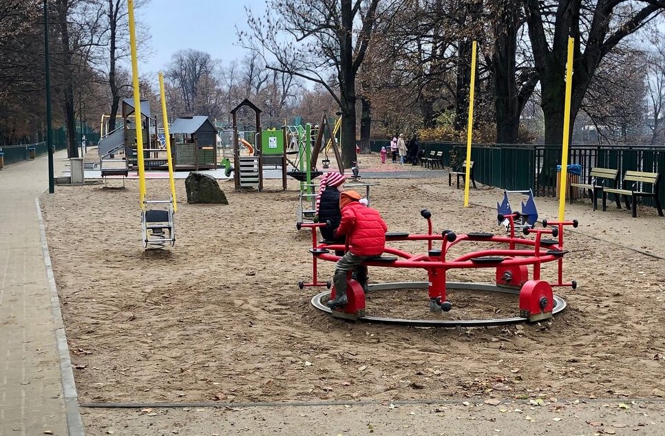 Two children in red jackets play on a circular red merry-go-round in a sandy playground lined with benches and surrounded by trees.