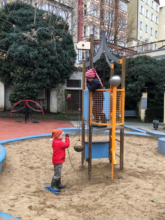 Two children play on a small sand-covered tower structure with pulleys and ropes in a city playground bordered by buildings and greenery.