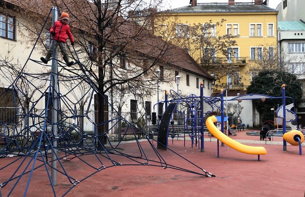 A child climbs a tall web of blue ropes in a compact city playground with yellow slides and play equipment, surrounded by colourful historic buildings in Prague.