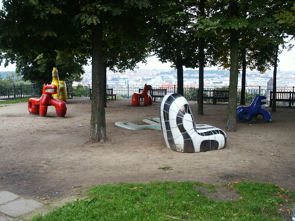 A shaded park area with whimsical, brightly colored sculptures shaped like animals and abstract forms beneath trees overlooking the city of Prague