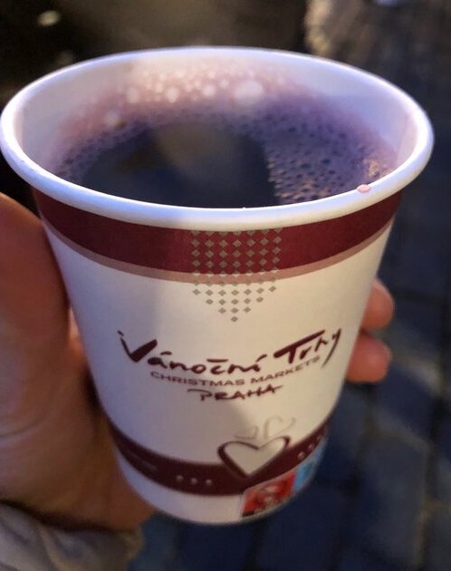 A hand holds a paper cup of hot mulled wine labeled “Vánoční trhy Christmas Markets Praha,” with steam rising from the drink.