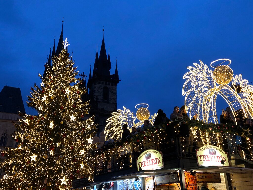 A beautifully lit Christmas tree and glowing archway decorations brighten a festive market square at dusk, with people gathered below and historic towers in the background.