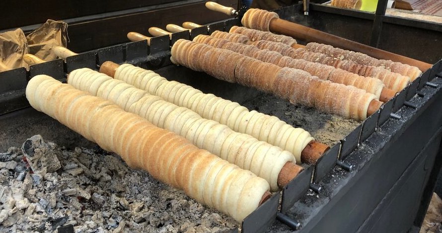 Rows of spiral pastries baking on wooden rods over hot coals at an outdoor Christmas market stall.