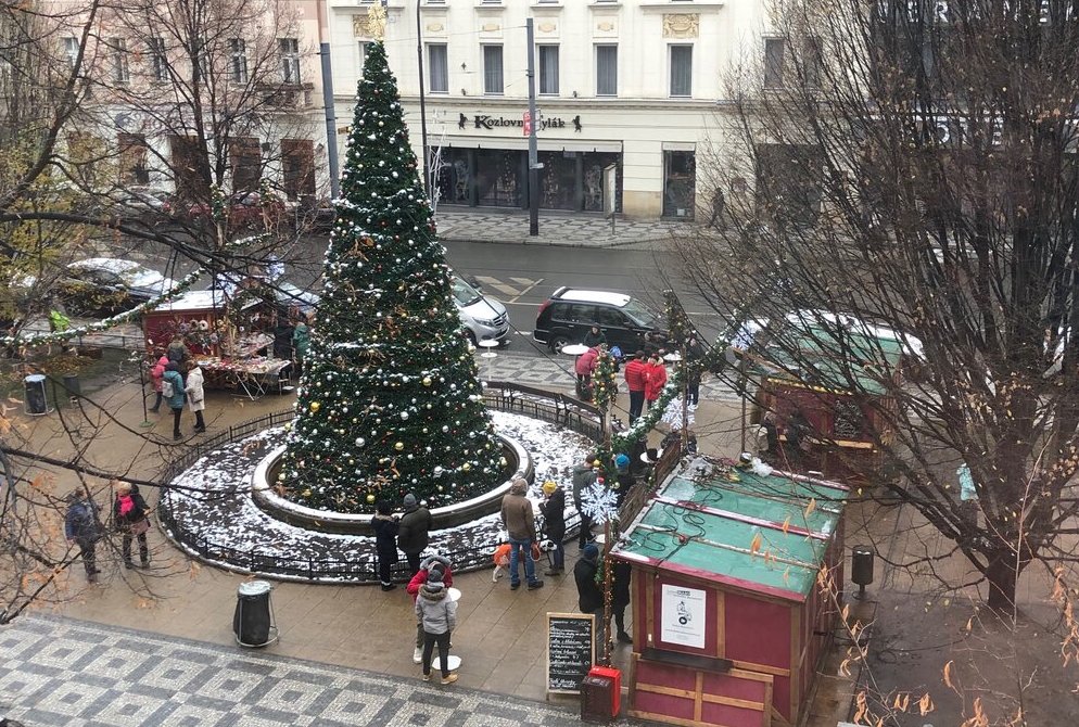 An overhead view of a small Christmas market with a large decorated tree at the center, surrounded by a few wooden stalls and visitors on a snowy street corner.
