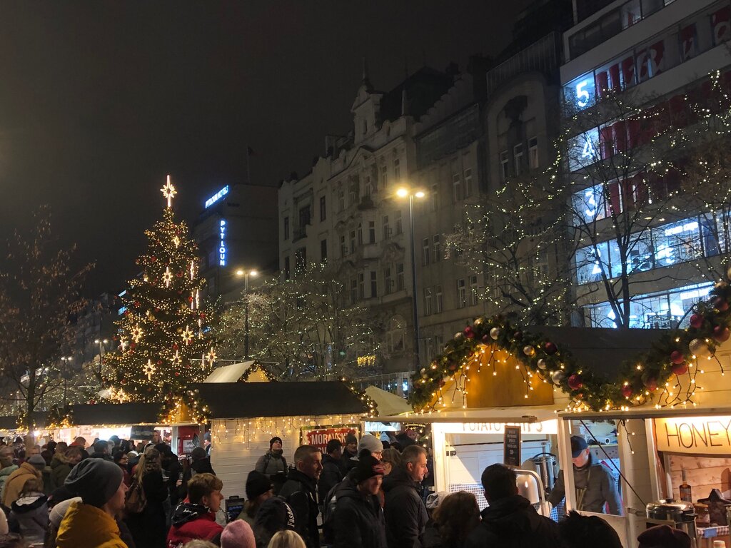 A bustling evening Christmas market with glowing stalls, a decorated tree, and festive lights strung through the trees along a city street.