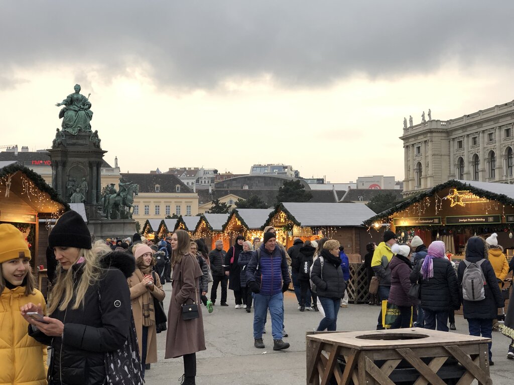 A busy Christmas market with garlanded huts and shoppers in winter coats is set against the backdrop of Vienna’s Maria Theresa statue and historic buildings under a cloudy sky.